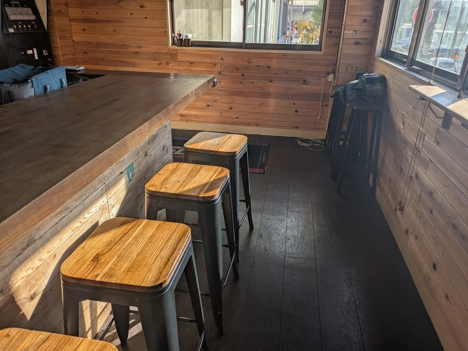 自然光が差し込む、木目調の壁とカウンターが特徴的なカフェまたはバーの店内。複数のハイスツールが並び、落ち着いた雰囲気を醸し出しています。