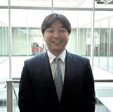 ビジネススーツを着たアジア人男性が、明るいオフィスのような場所で笑顔を見せているポートレート