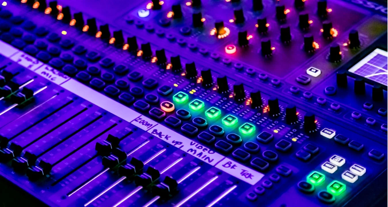 紫と緑の光で照らされたオーディオミキサーのクローズアップ画像。多くのフェーダー、ノブ、ボタンがあり、ライブイベントや音楽制作で使用されるプロフェッショナルな音響機器です。