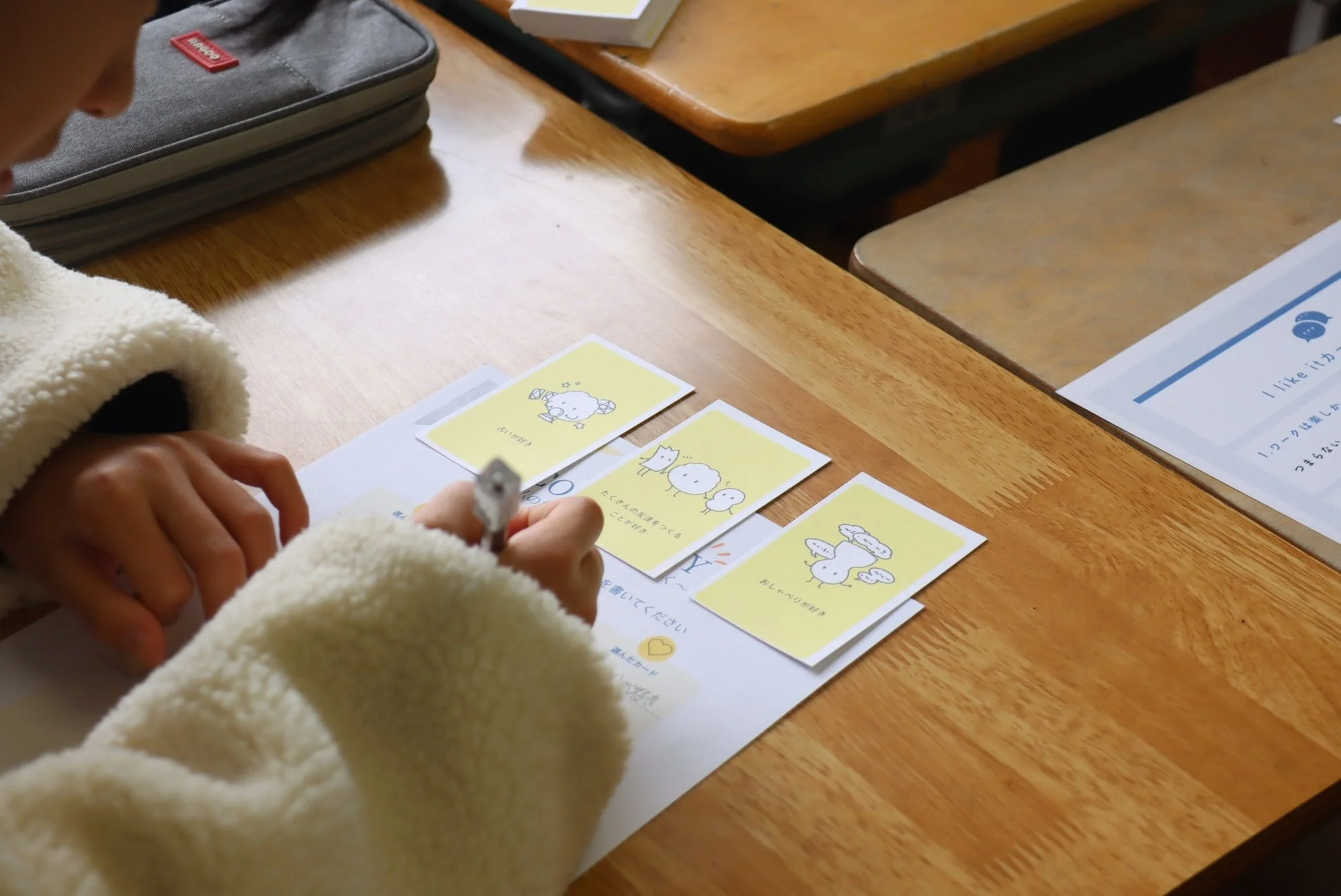 教室で、子供が黄色のイラスト入りカードを見ながらワークシートに書き込んでいる様子