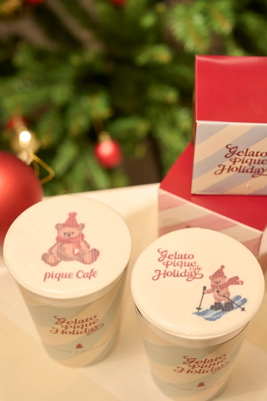 クリスマスツリーを背景に、ジェラートピケホリデーのカップとギフトボックスが写っています。カップの蓋には可愛らしいテディベアのイラストが描かれており、ホリデーシーズンの贈り物やカフェタイムを連想させます。