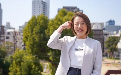 白いTシャツと薄い色のジャケットを着用した女性が、屋外の都市景観を背景に笑顔で立っている