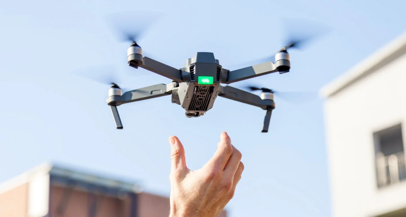 青い空を背景に、一人の手がホバリングしているドローンに手を伸ばしている瞬間を捉えた画像です。ドローンは飛行中で、プロペラが回転しています。