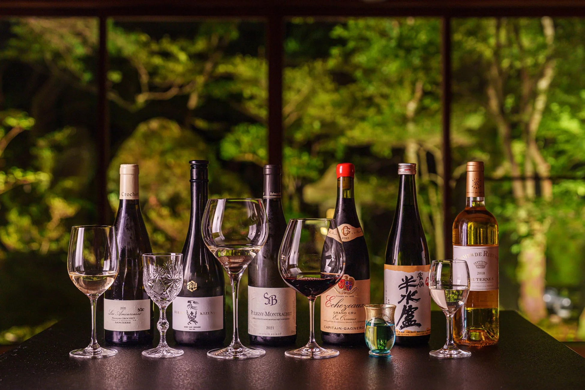 ワインボトルとグラスが並べられたテーブル、背景には緑豊かな庭園