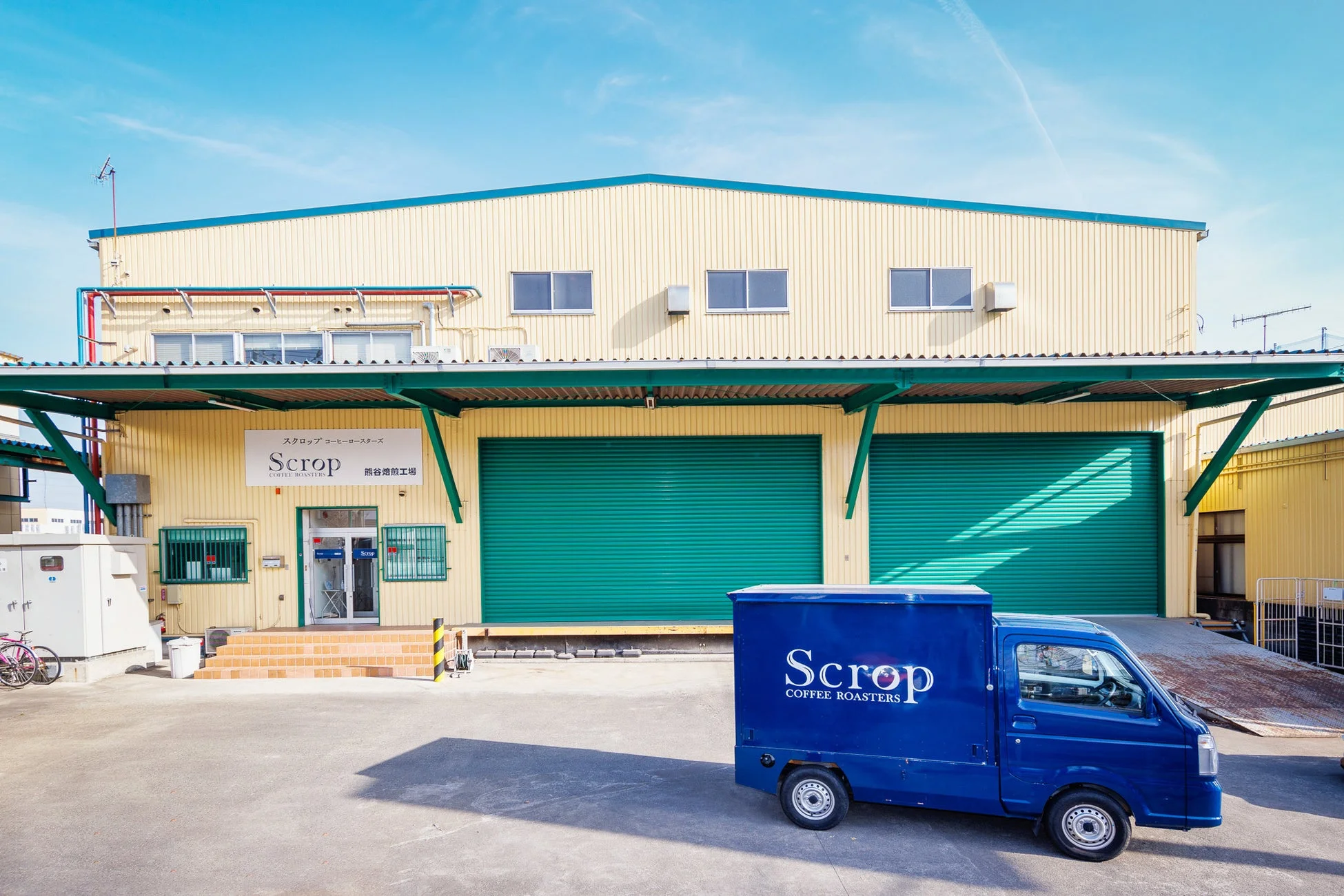 「Scrop COFFEE ROASTERS 熊谷焙煎工場」と書かれた黄色の建物と、同社のロゴが入った青い配送トラックが写っています。晴れた日の工場外観です。