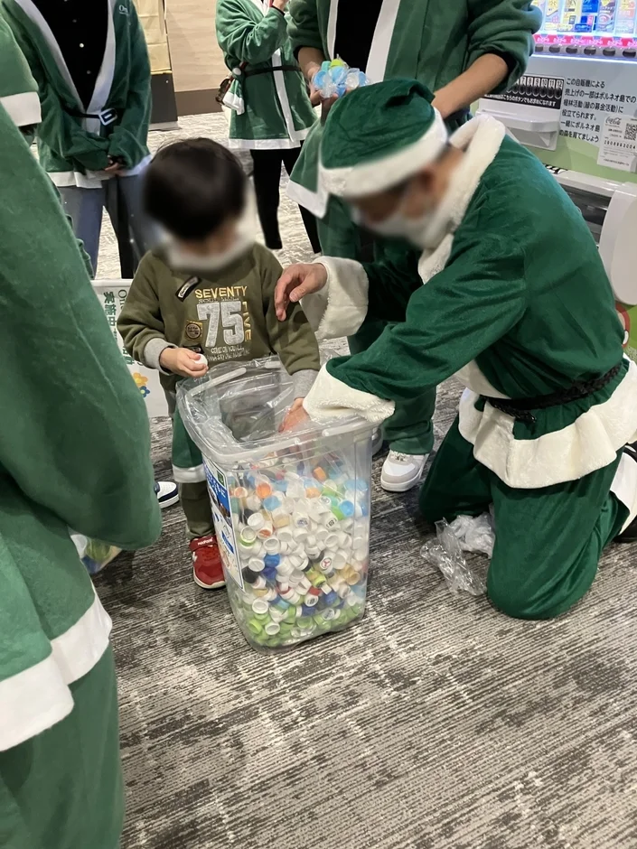 緑色のサンタ衣装を着た大人と子供が、ペットボトルキャップの回収活動を行っている様子。透明な容器には色とりどりのキャップがぎっしり詰まっており、チャリティやリサイクルを目的としたイベントの一環と見られる。