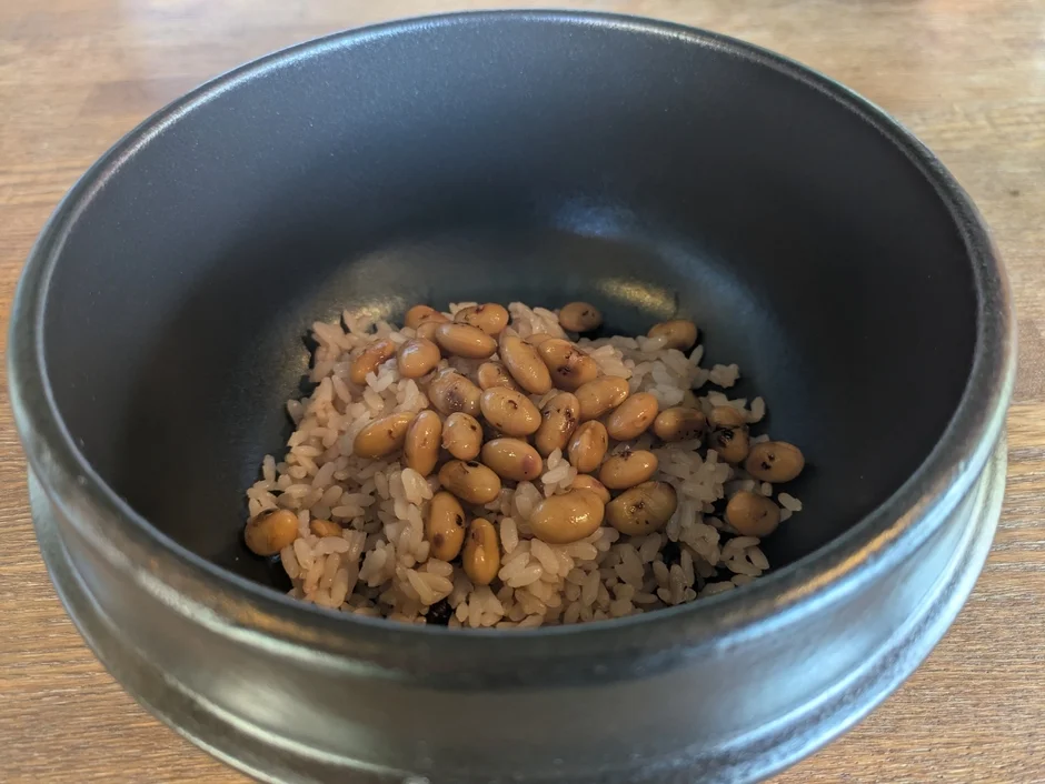 黒い器に入ったご飯と豆の料理です。健康的な和食の一品で、ご飯の上にたっぷりの煮豆がのっています。