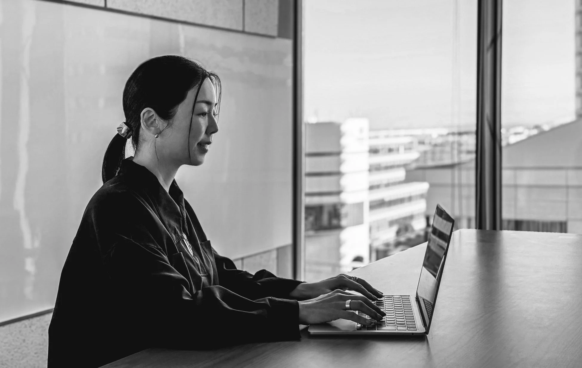 オフィスのような空間で窓の外を背景にノートパソコンで作業する女性の横顔