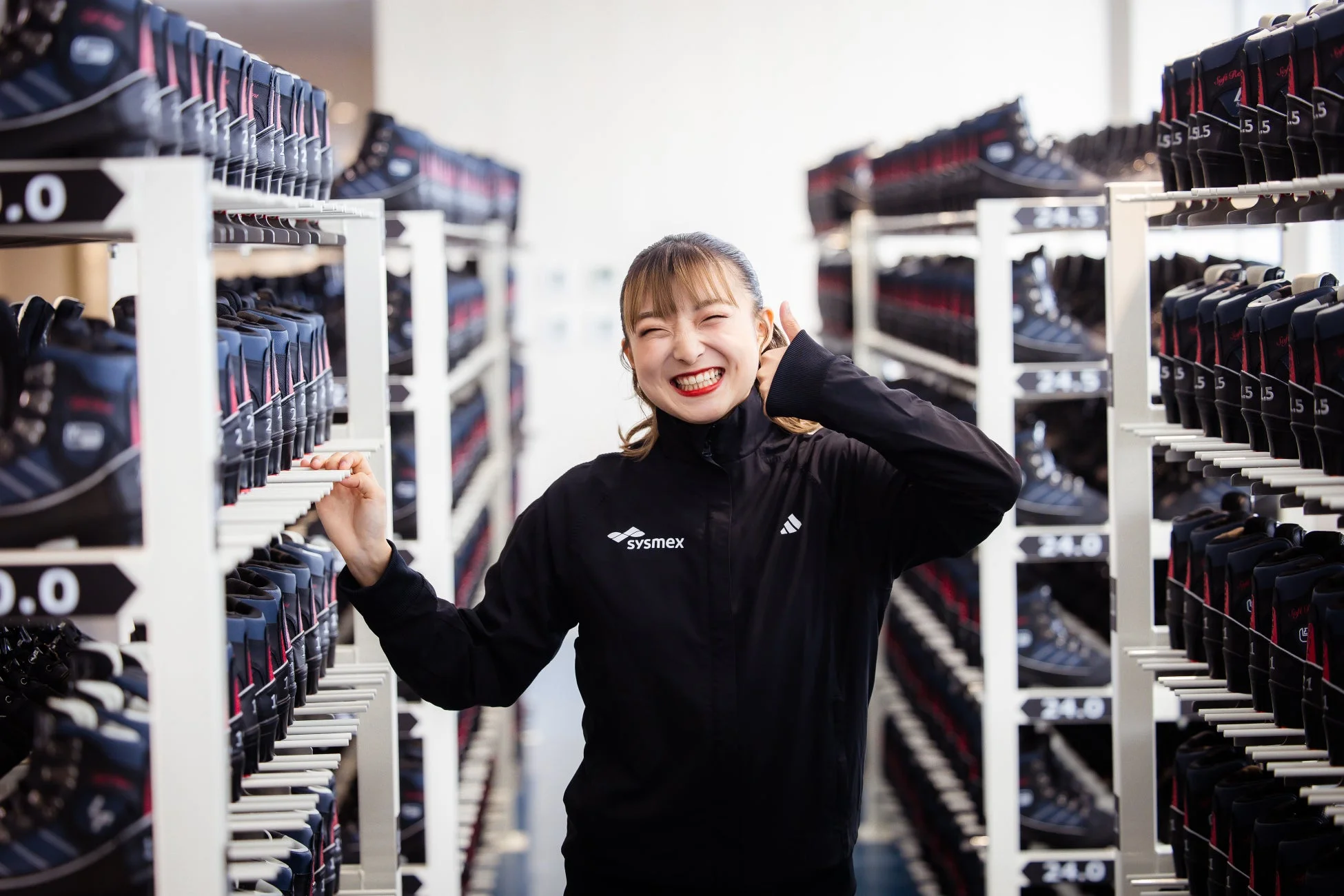 アイススケートがずらりと並んだ棚の間で、黒いジャケットを着た女性が満面の笑みを浮かべている。