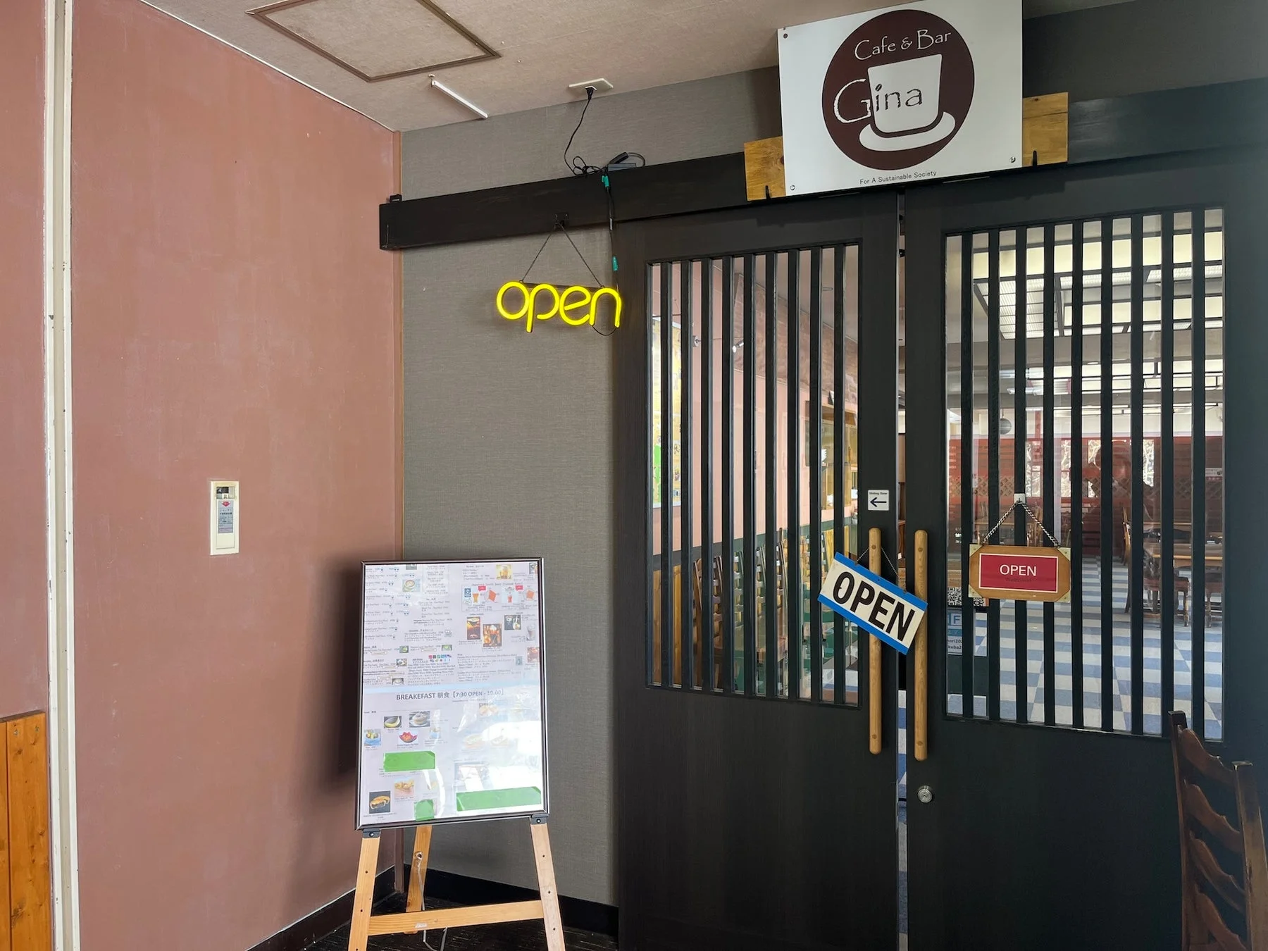 「Cafe & Bar Gina」の入り口の様子。ネオンの「open」サインや複数の「OPEN」の看板が目立つ。店内は営業中で、入り口横には朝食メニューの看板が置かれている。