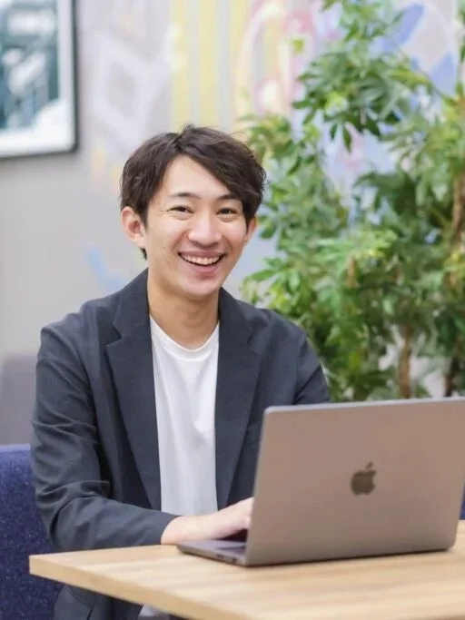 笑顔でカメラを見つめる若いアジア人男性が、カジュアルなジャケット姿でノートパソコンを使用している様子