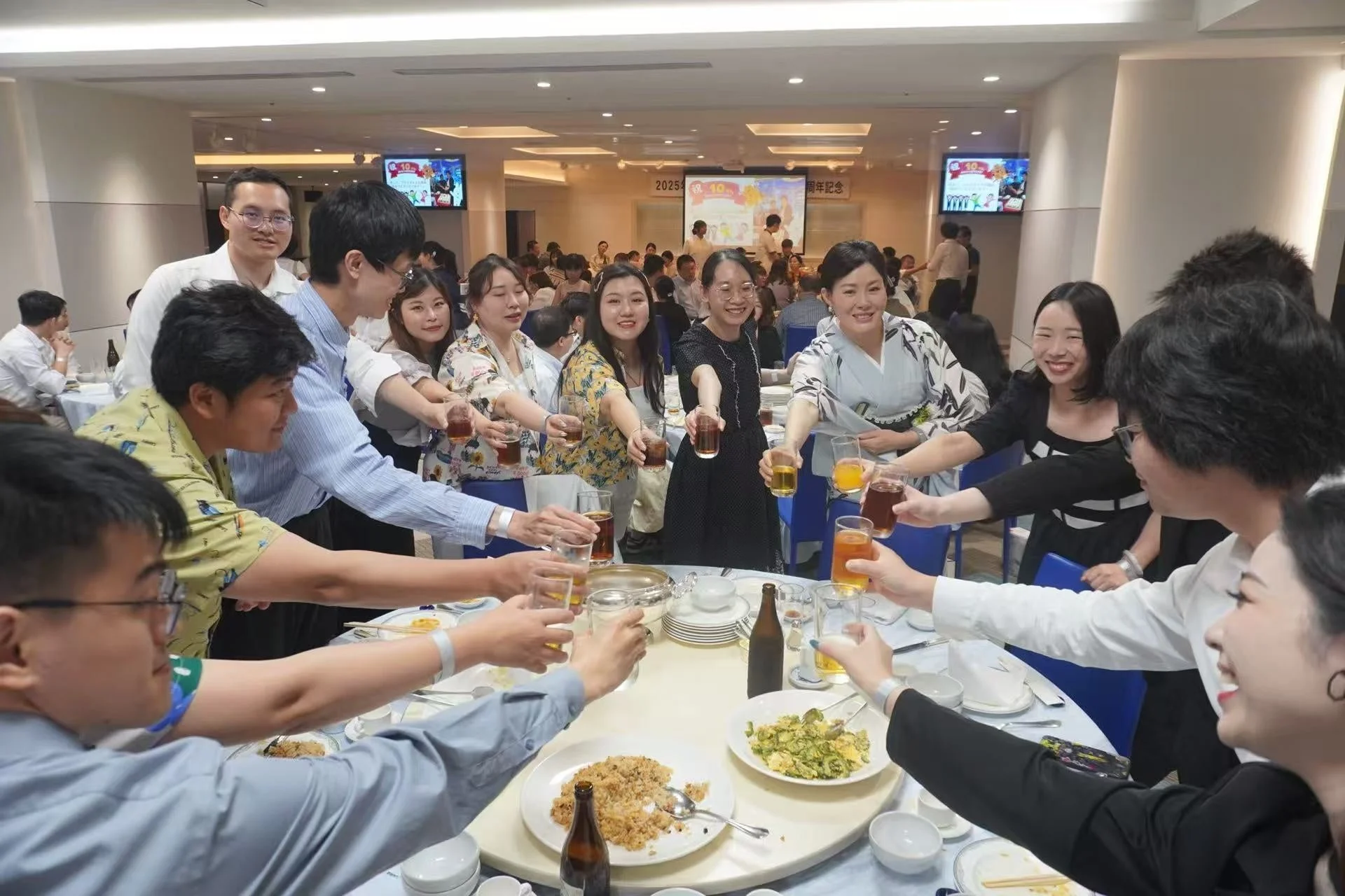 大勢の人々が円卓を囲み、笑顔で乾杯をしている賑やかな宴会の様子です。周年記念のような祝賀イベントで、参加者たちは飲み物や食事を楽しみながら交流を深めています。