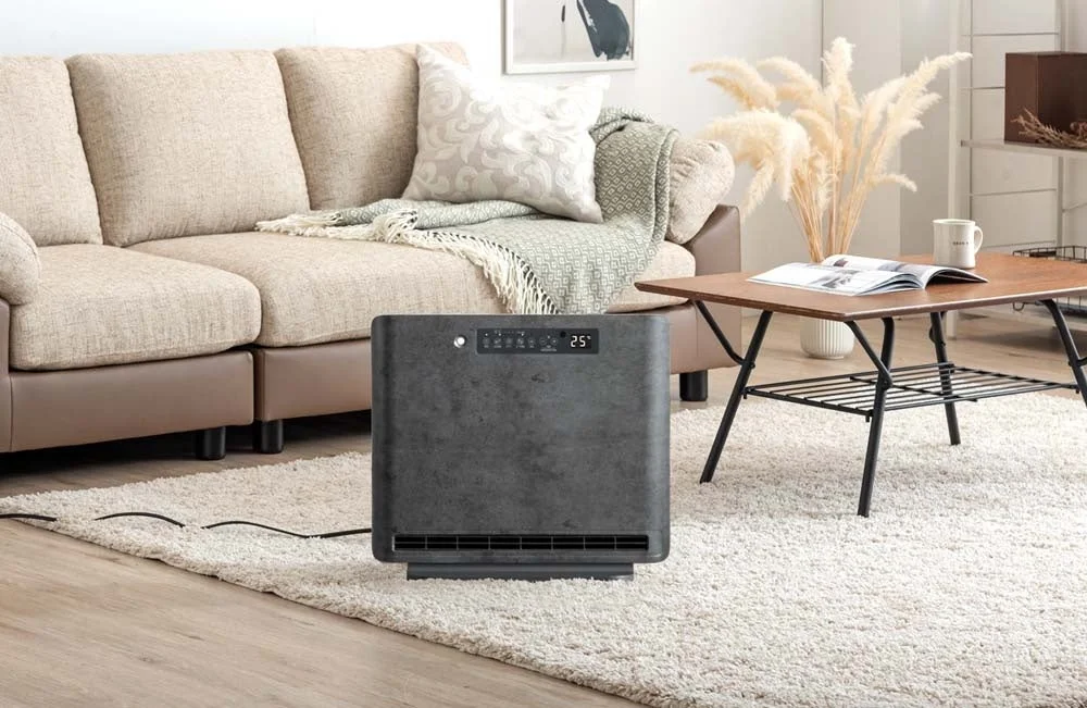 ベージュのソファが置かれたリビングルームに、スタイリッシュなダークグレーの暖房器具が白いふわふわのラグの上に設置されています。木製のコーヒーテーブルには雑誌とマグカップがあり、全体的に落ち着いたモダンな雰囲気です。