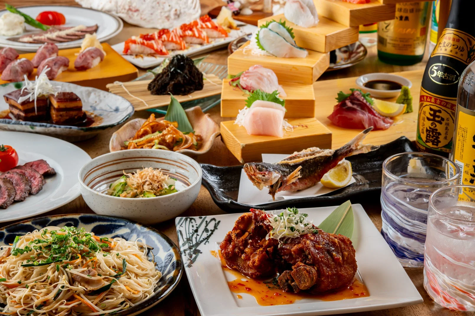 刺身、寿司、肉料理、魚料理、麺類など、多種多様な和食が豪華に並べられた食卓の風景。沖縄の泡盛も添えられており、賑やかな宴の雰囲気を伝えています。