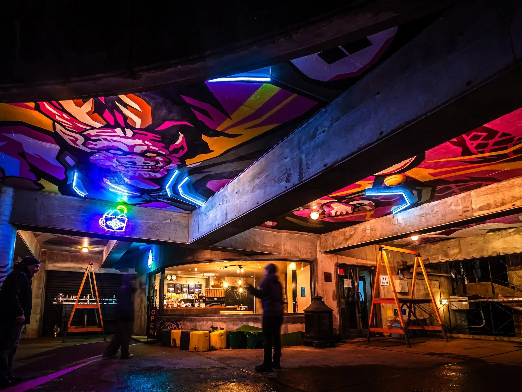 ネオンライトとグラフィティアートで彩られた都市の屋内空間。天井には鮮やかな壁画と青いネオンが輝き、奥にはカフェが見えます。夜の活気ある雰囲気を捉えた一枚です。