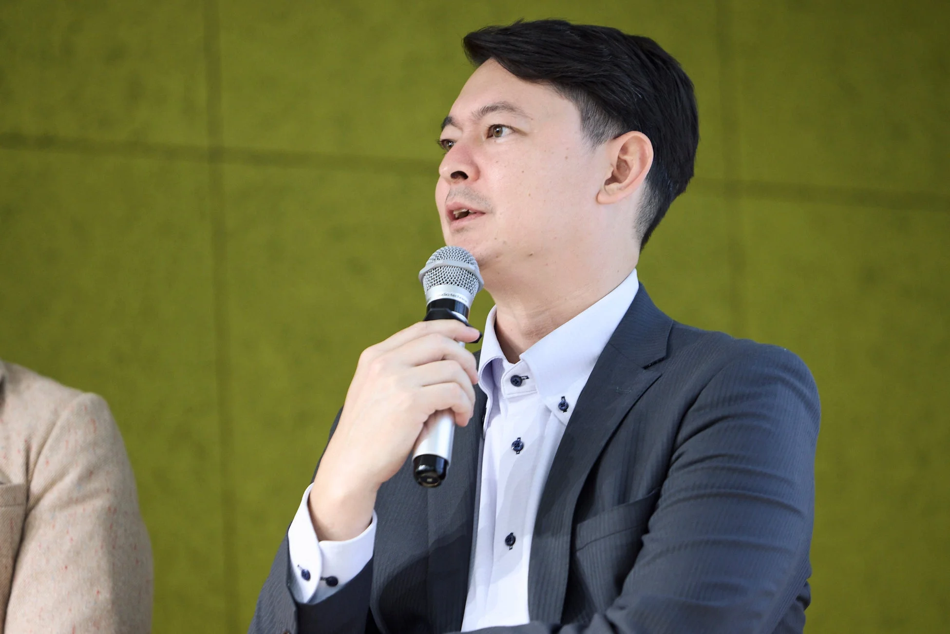 マイクを手に持ち、真剣な表情で話すアジア人男性