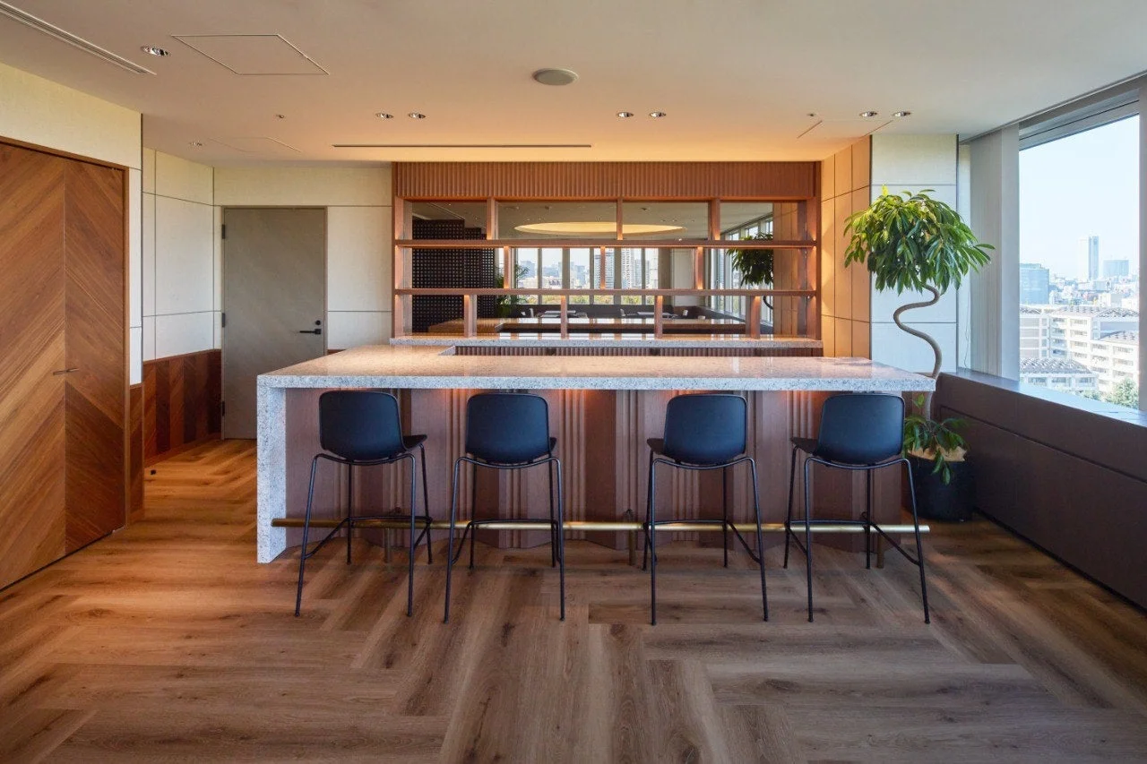 モダンな空間に石材のカウンターバーと4脚のバースツールが配置されています。ヘリンボーン柄の木製フローリングと木製パネルが特徴的で、窓からは都市の景色が広がり、洗練された雰囲気を演出しています。