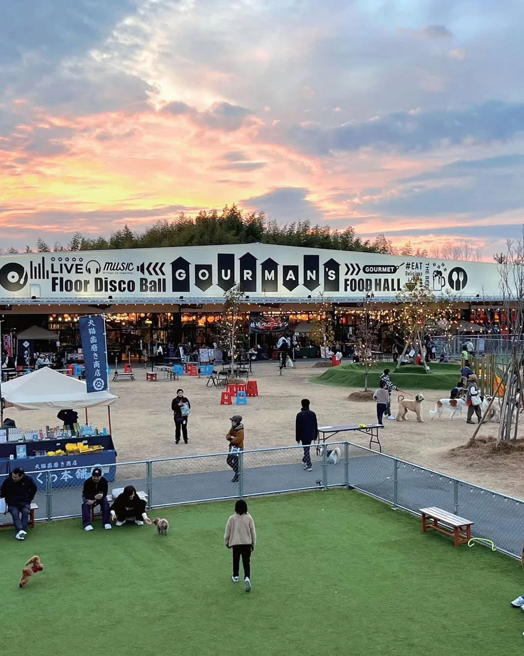 夕焼けの空の下、屋外のフードホール「GOURMAN'S FOOD HALL」で人々が犬とくつろいでいる様子。ドッグランや様々な店舗があり、活気ある夕暮れの光景です。