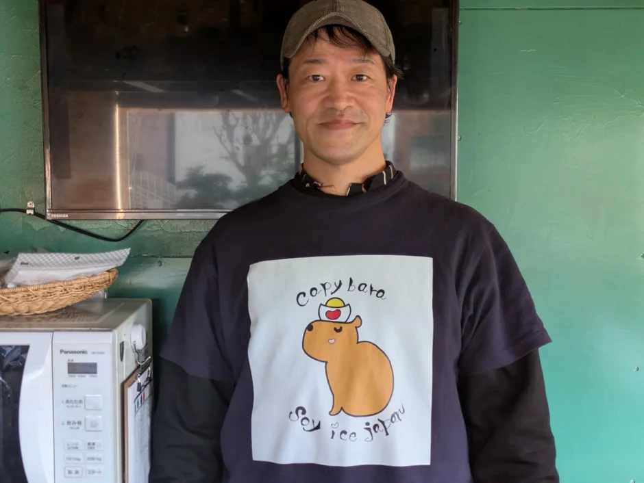 カピバラのイラストと「soy rice japan」の文字がプリントされたTシャツを着た男性が写っています。背景には電子レンジとテレビが見えます。
