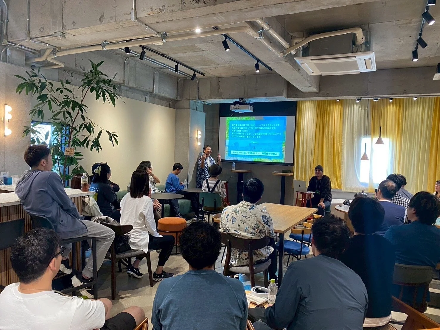 モダンな空間でプレゼンテーションが行われています。一人の話し手がプロジェクターに資料を映し出し、多くの参加者が熱心に耳を傾けています。