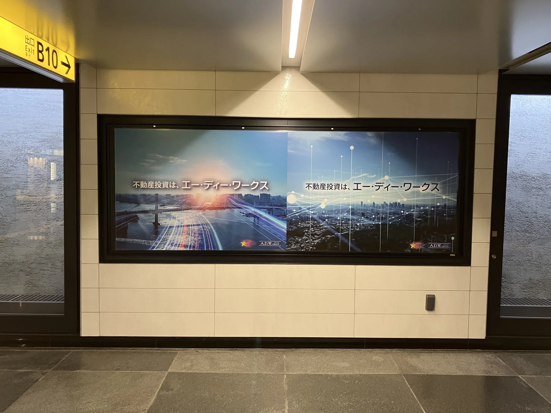 地下通路のような場所に設置された、不動産投資会社「エーディーワークス」の広告です。広告には、光の軌跡やネットワークが重なる都市の風景が描かれています。横には「出口 B10」の案内表示が見えます。
