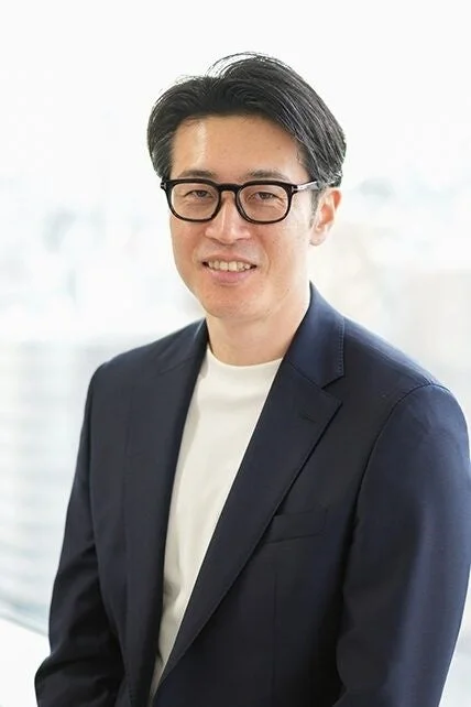 眼鏡をかけた東洋系の男性が、紺色のジャケットと白いTシャツを着てにこやかにカメラを見つめているポートレート写真です。