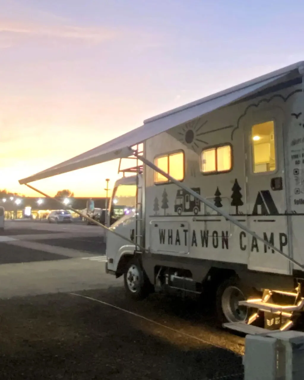 夕焼け空の下、キャンピングカーが駐車場に停まっています。日よけのタープが広げられ、車内には明かりが灯り、「WHATAWON CAMP」の文字が見えます。