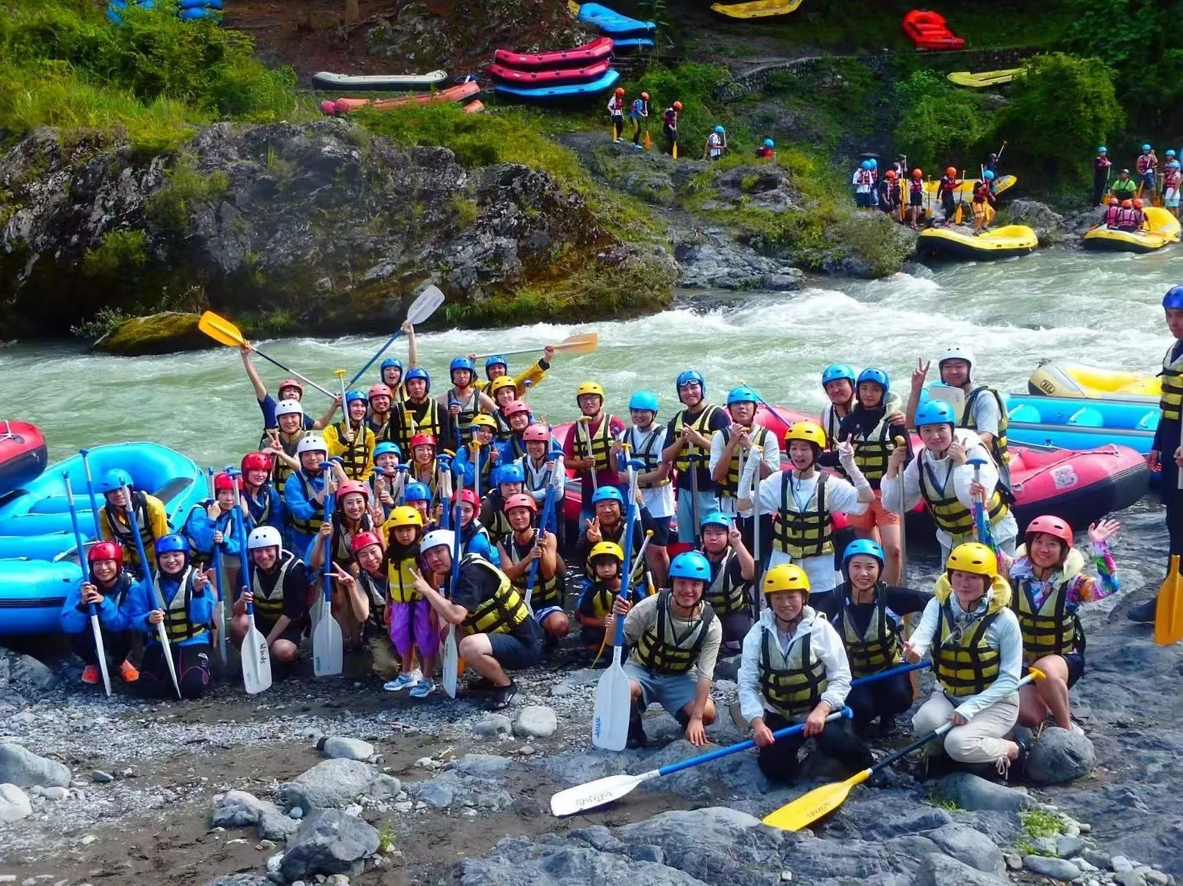 川辺でヘルメットとライフジャケットを着用した大勢のグループが、パドルを持って笑顔で集合写真を撮っています。ラフティング体験の前か後で、楽しそうな雰囲気が伝わります。背景には複数のゴムボートと緑豊かな自然が広がっています。