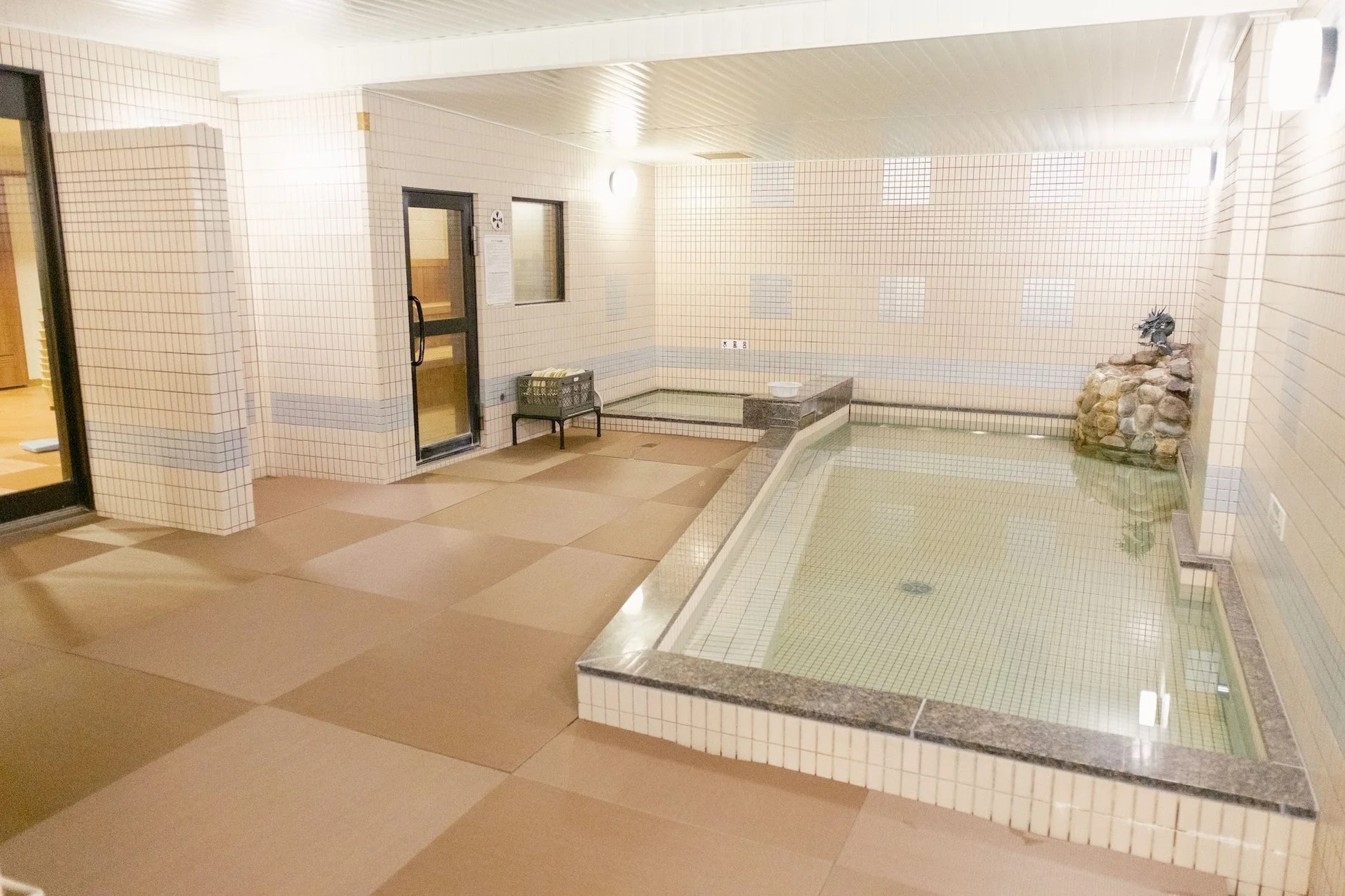 旅館やホテルにある清潔感のある大浴場の様子です。白いタイル張りの空間に、大小二つの浴槽があり、利用者がリラックスできる環境が整っています。浴槽の脇には石積みの上に龍の置物が配置され、和風の趣を加えています。