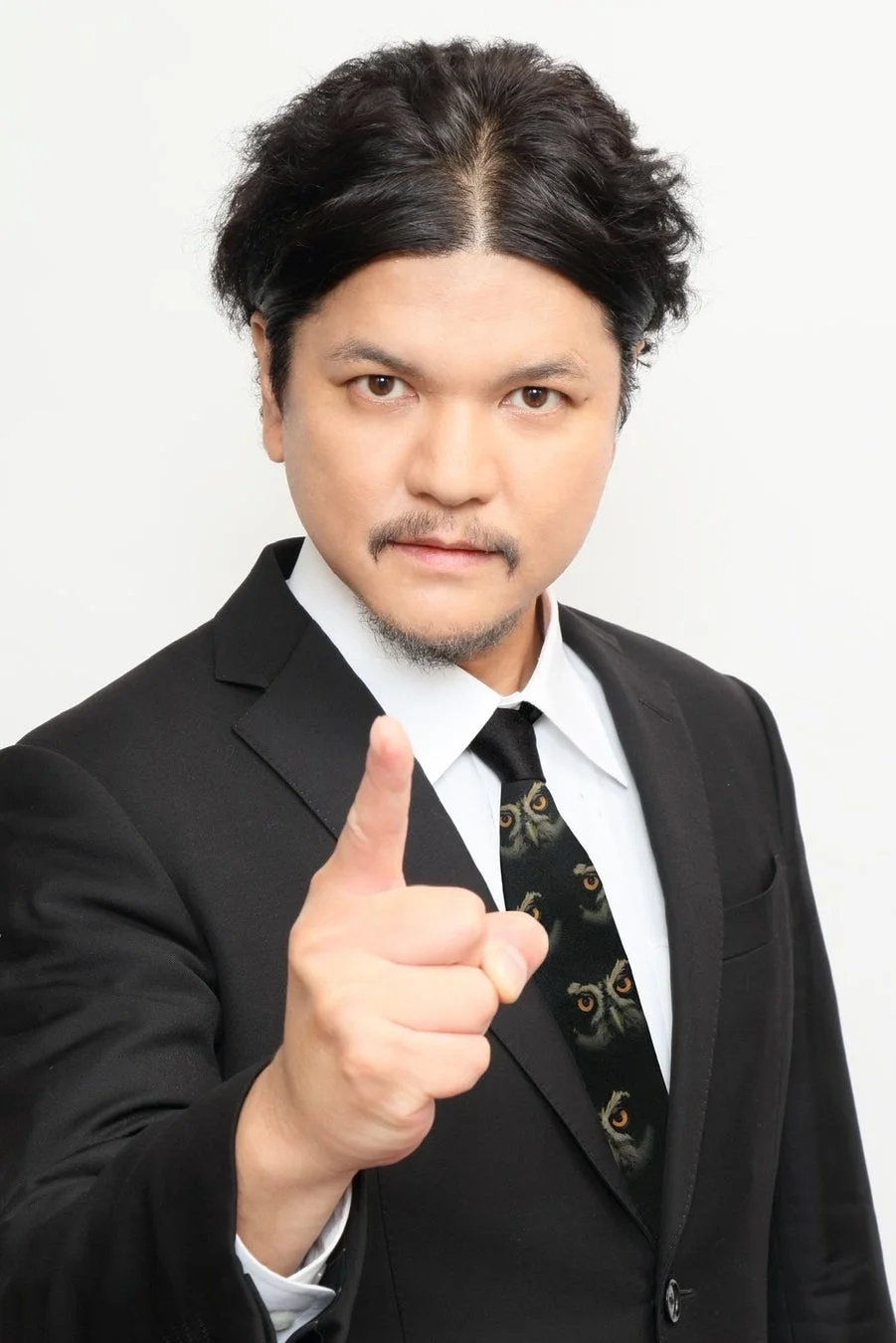 スーツを着た男性が、正面に向かって指をさしています。ひげを生やし、フクロウの顔がデザインされたユニークなネクタイを着用しており、真剣な表情をしています。