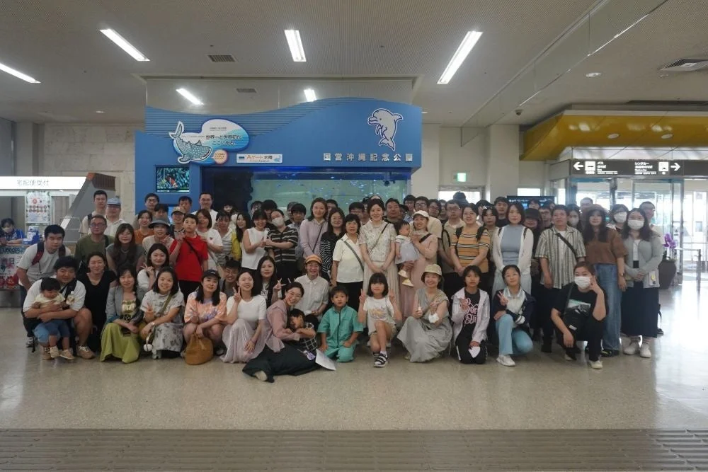 国営沖縄記念公園内の美ら海水族館と思われる場所で、大人数で記念撮影をしている様子です。多くの家族連れが笑顔でポーズをとっており、和やかな旅行の雰囲気が伝わります。