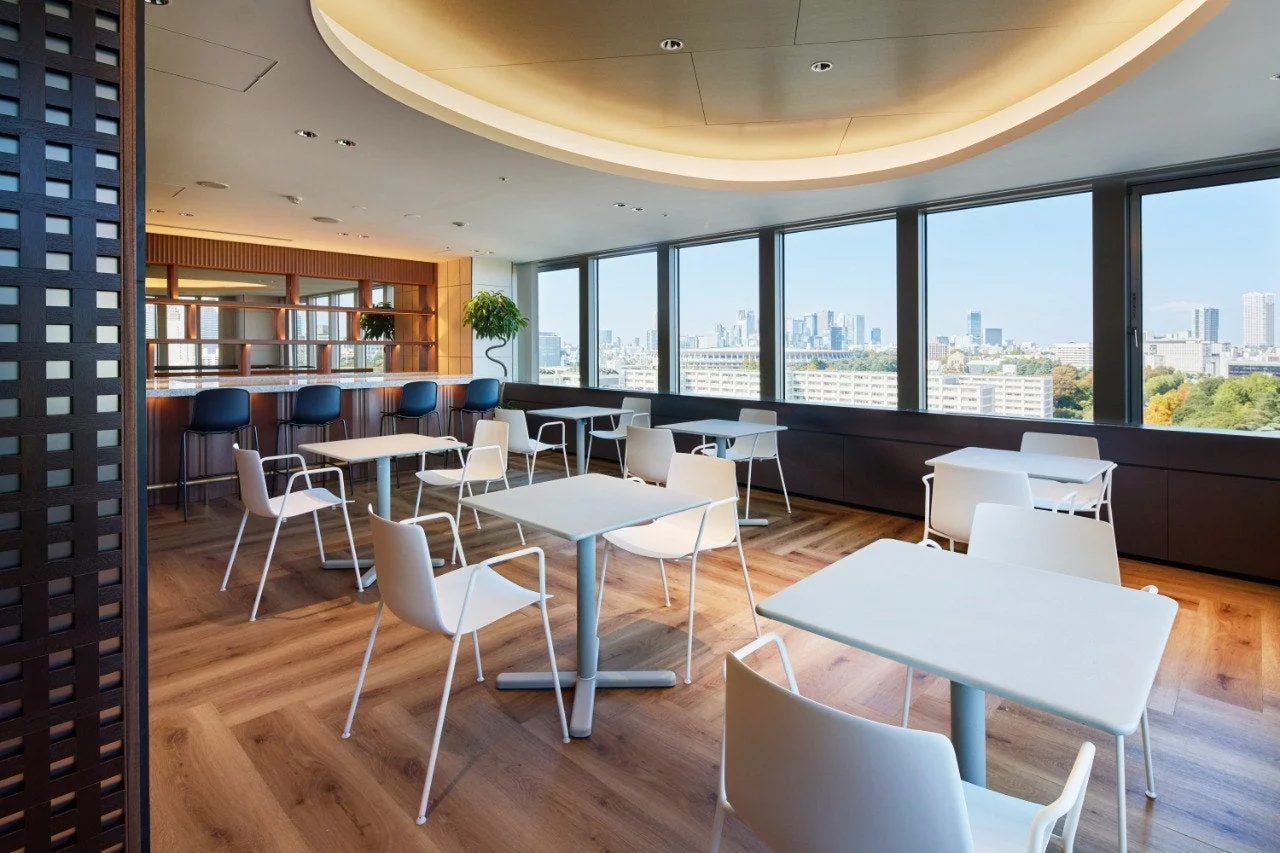 都市のスカイラインを一望できる明るくモダンなオフィスラウンジまたはカフェスペース。バーカウンターと複数のテーブル席が配置され、木目調の床と白い家具が清潔感のある落ち着いた空間を演出しています。