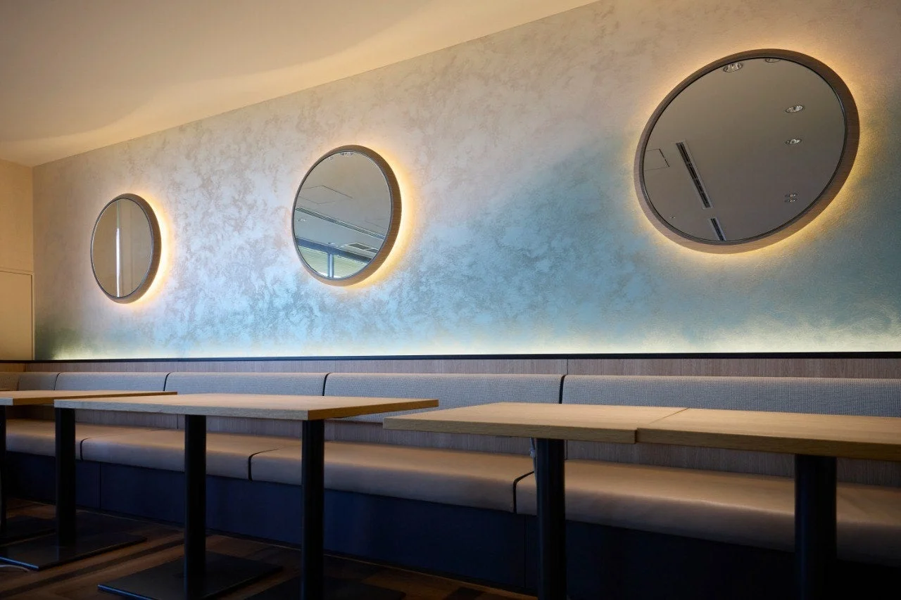 モダンなカフェまたはレストランの店内。テクスチャのある壁にはバックライト付きの丸い鏡が複数飾られ、その下にはベンチシートと木製のテーブルが並んでいます。落ち着いた雰囲気のインテリアデザインです。