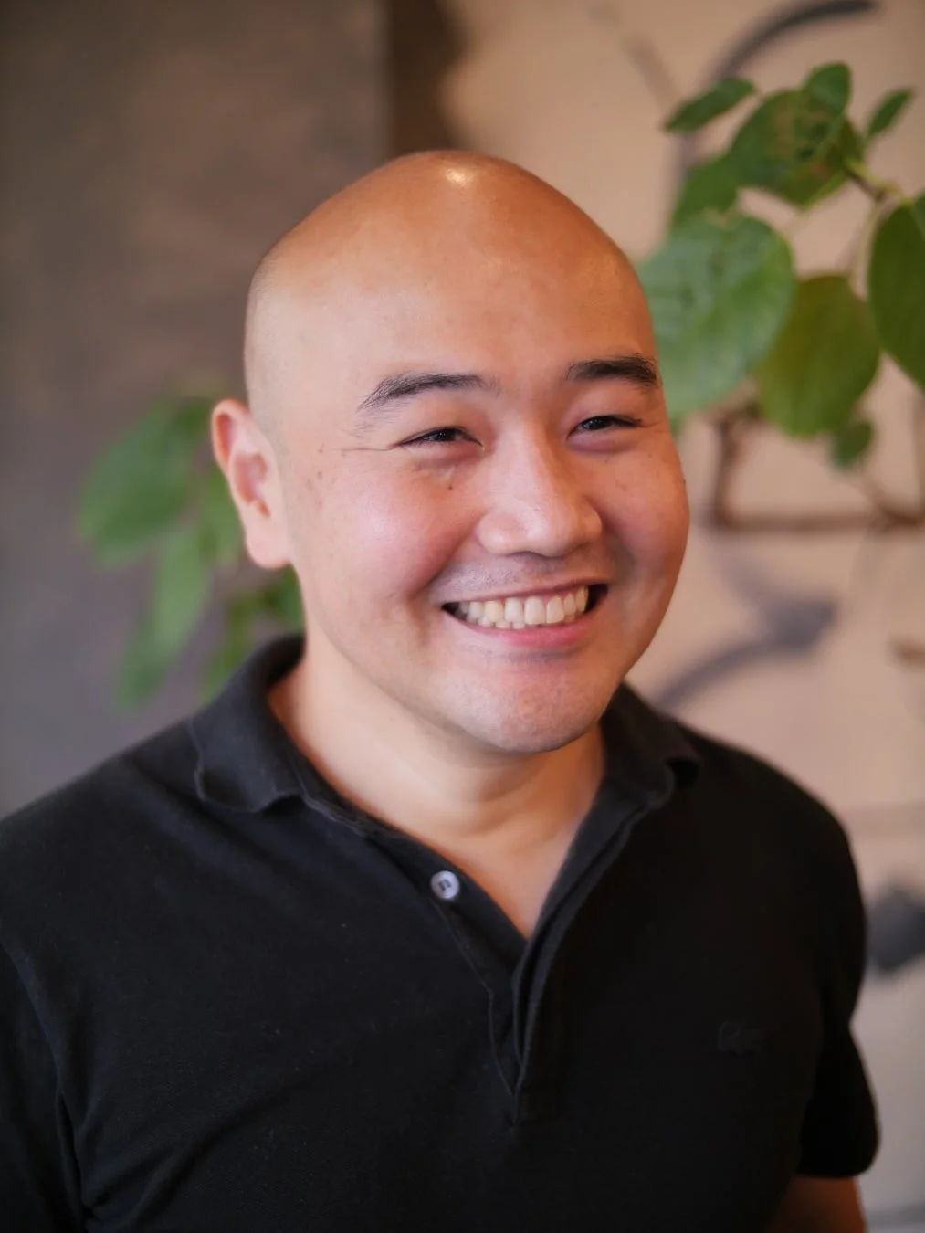 黒いポロシャツを着た坊主頭のアジア人男性が、明るい笑顔を見せています。背景はぼやけており、屋内でのポートレート写真です。