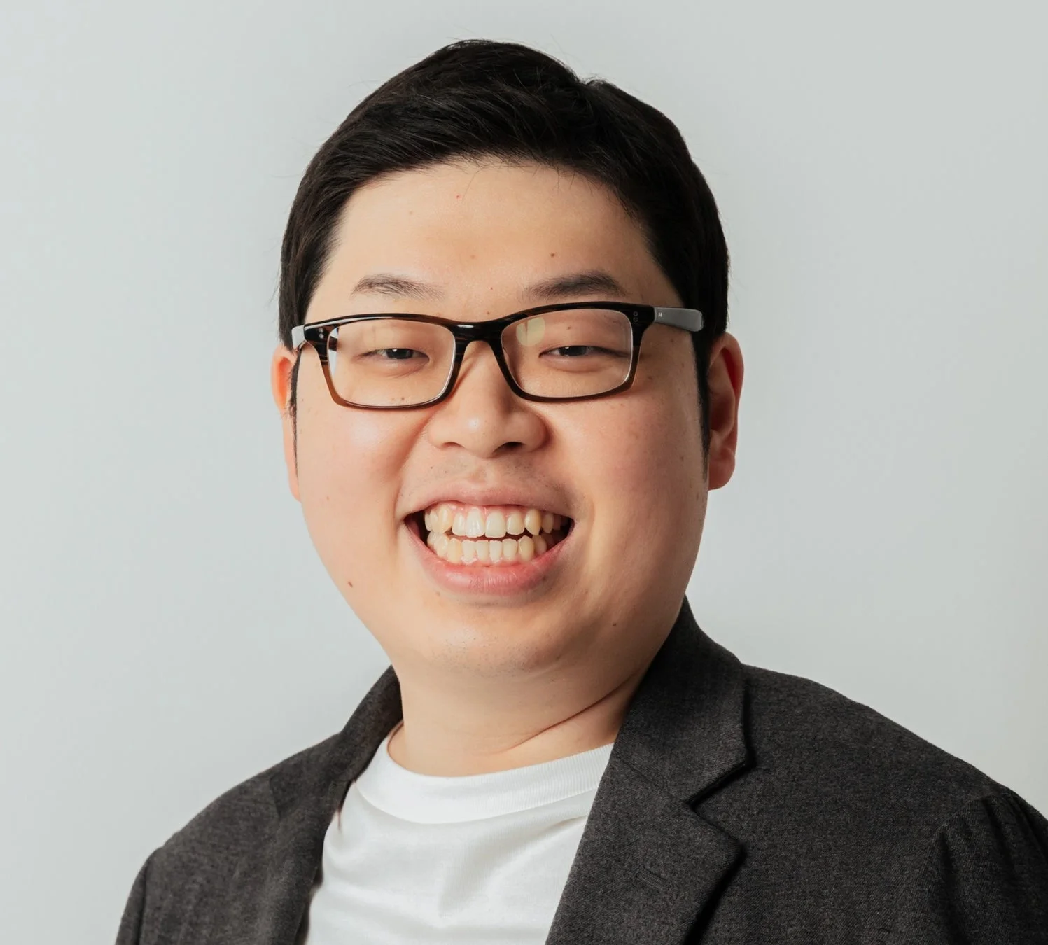 白い背景の前で、眼鏡をかけたアジア人男性が満面の笑みを浮かべているポートレート写真です。黒いジャケットと白いTシャツを着用しており、親しみやすい印象を与えます。