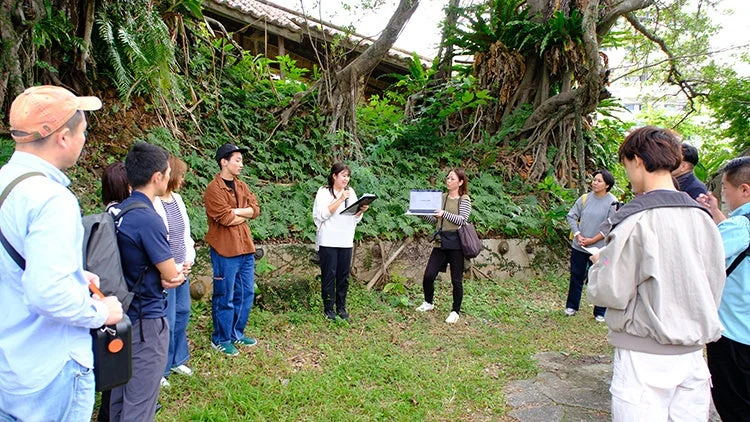 緑豊かな屋外で、数人の参加者が集まり、2人の女性が説明やプレゼンテーションを行っている様子