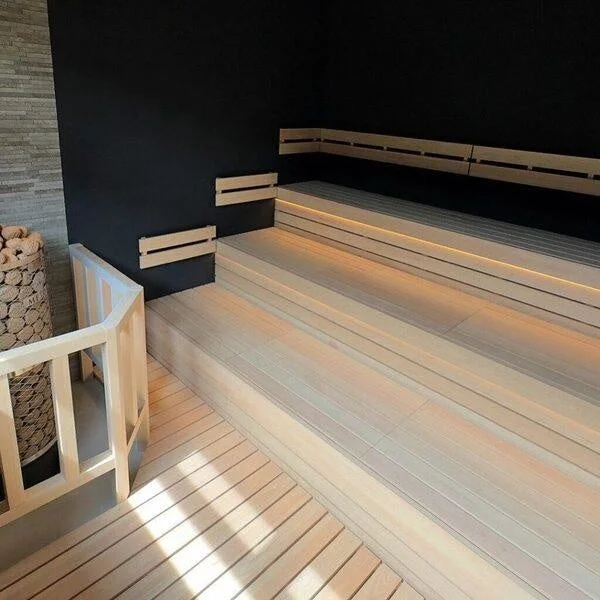 モダンで洗練されたデザインのサウナ室の内部。明るい木製の多段ベンチと、コントラストをなす黒い壁が特徴。ベンチ下には間接照明が施され、リラックスできる空間を演出。左手には石積みのサウナヒーターが見える。