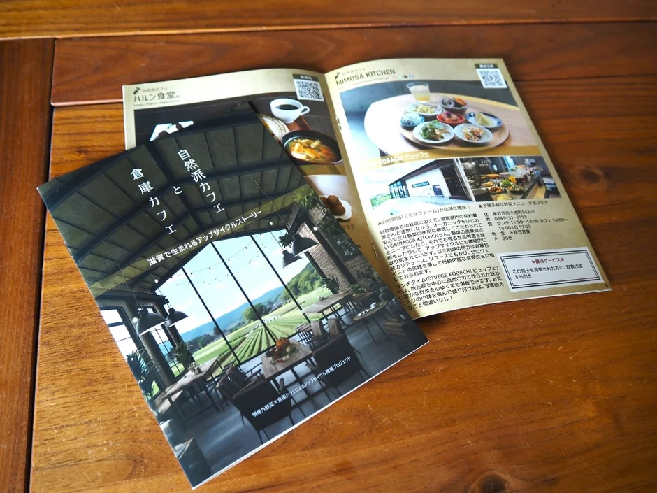 木製テーブルに、自然派カフェ「倉庫カフェ」と「MIMOSA KITCHEN」のパンフレット。開放的な空間のカフェ、滋賀野菜ビュッフェのMIMOSA KITCHENの店舗情報や、アップサイクルへの取り組みが紹介されています。