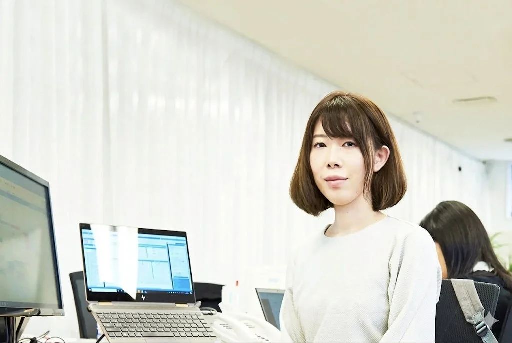オフィスで働く若い日本人女性が写っています。彼女はデスクに座り、ノートパソコンとモニターが置かれた職場で、穏やかな表情でカメラを見ています。