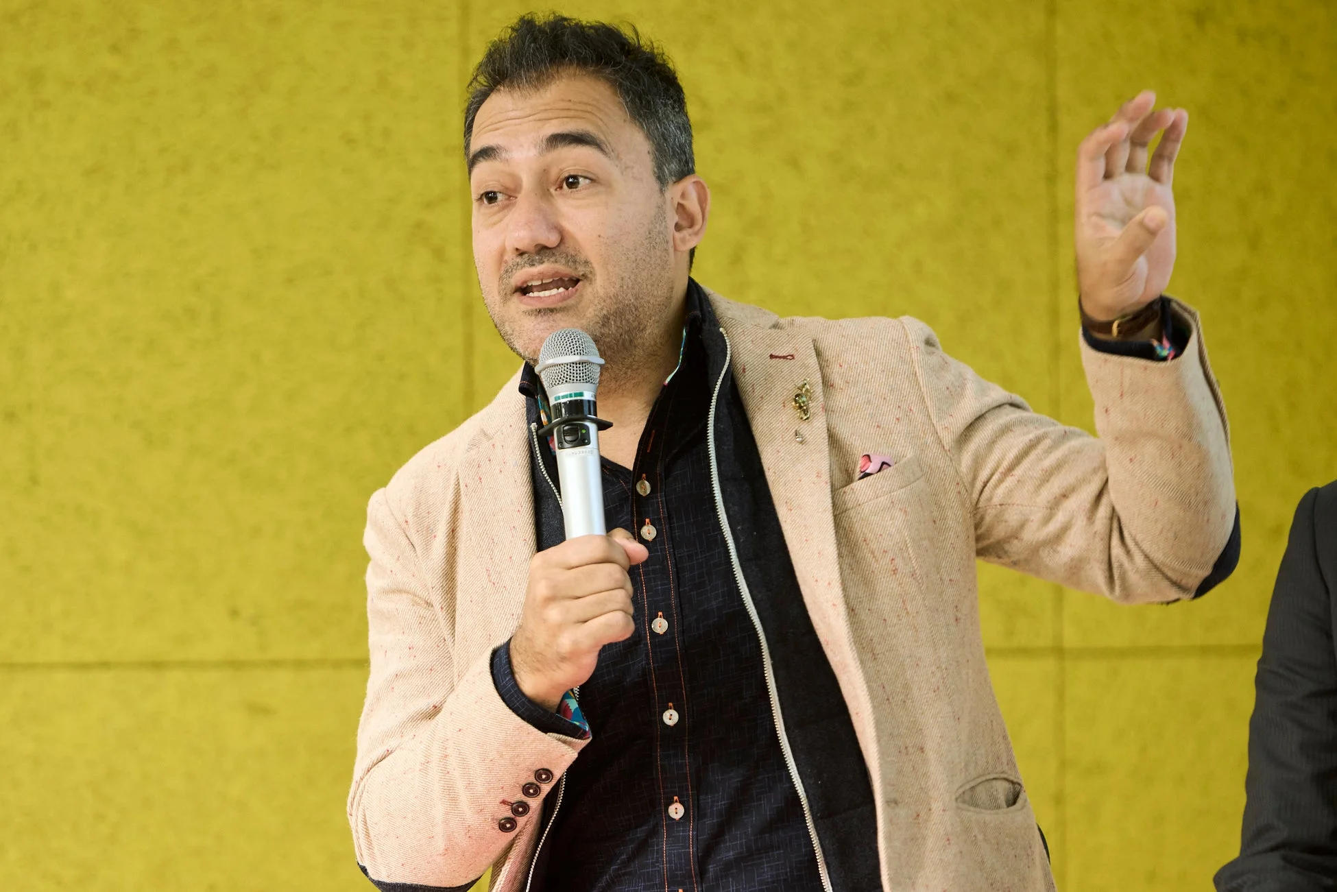黄色の背景の前で、マイクを片手に身振り手振りを交えながら話す男性