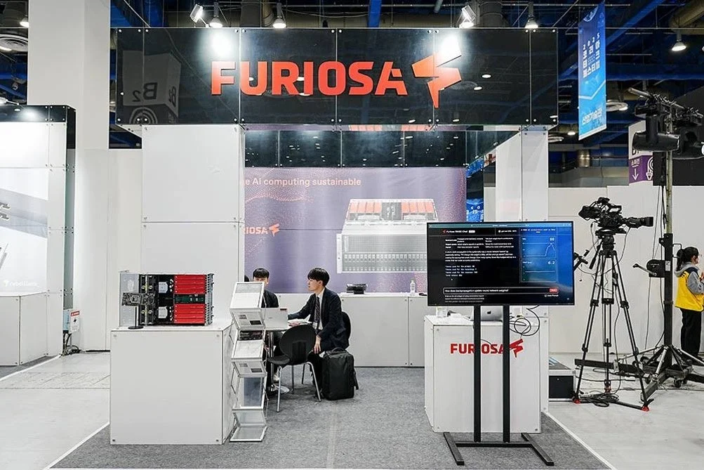 フリオサAIの展示ブースで、AIコンピューティングソリューションが紹介されています。来場者が技術的な説明を受け、モニターにはAIに関する情報が表示されており、活気あるイベントの様子がうかがえます。