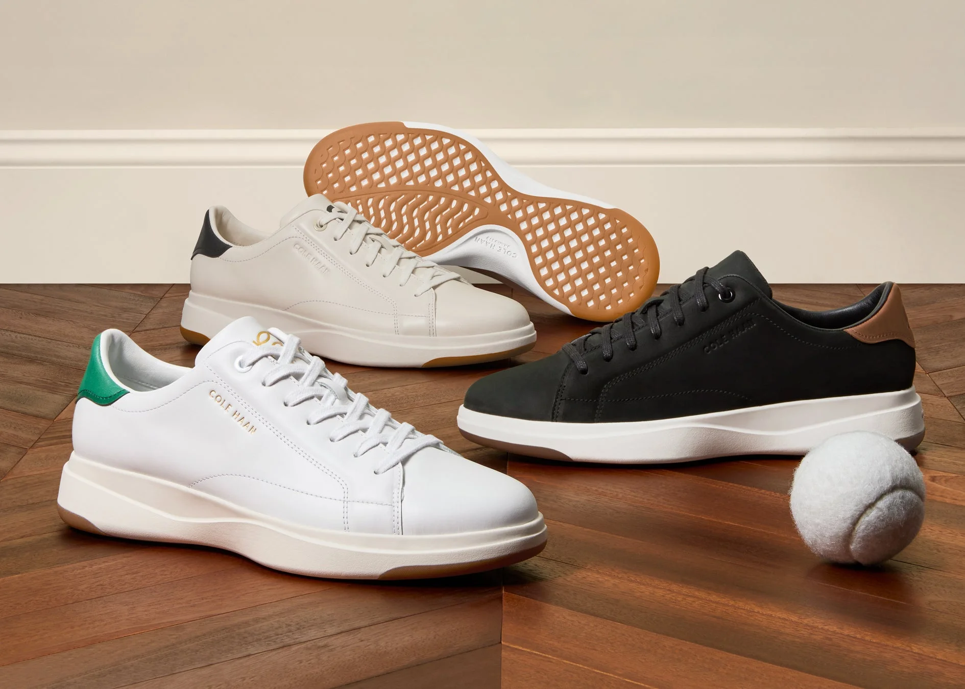 木製の床に3足のコールハーンのスニーカーが置かれ、テニスボールも添えられています。白、ベージュ、黒の異なる色のシューズがスタイリッシュに並べられています。