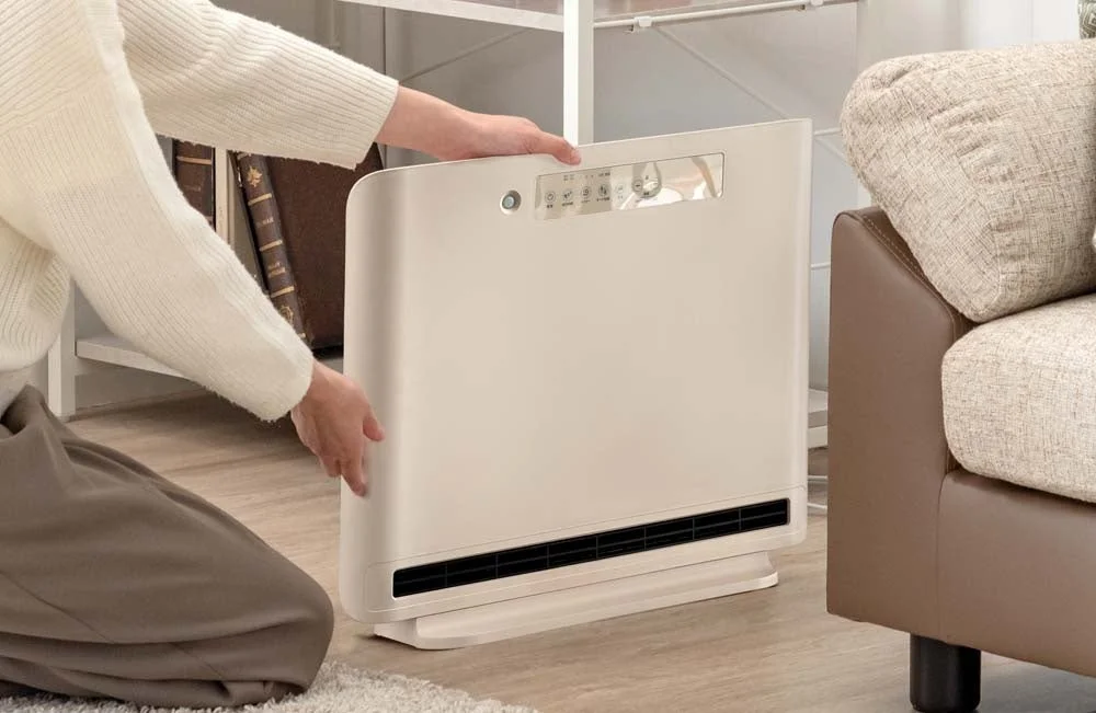 リビングでベージュ色の多機能家電（暖房、送風、加湿など）を床に置いている女性の様子。部屋を快適にするための準備をしている瞬間を捉えた一枚です。