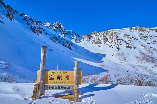 中央アルプス、宝剣岳の千畳敷カールが広がる冬の雄大な景色です。真っ青な空の下、雪に覆われた山々と広大な雪原が広がり、景勝地の標識が設置されています。
