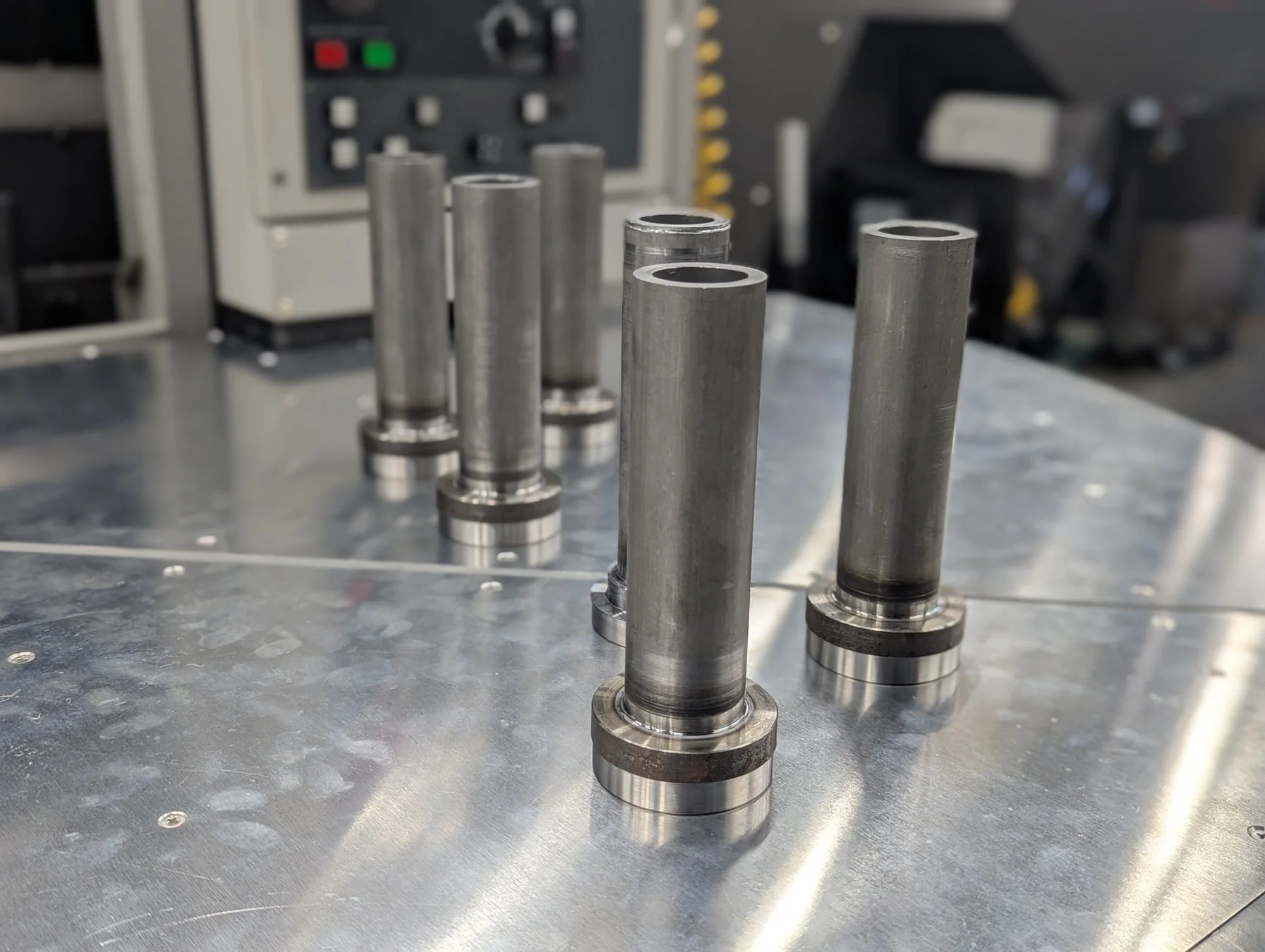銀色の作業台に、底にフランジが付いた金属製の円筒形部品が複数並べられています