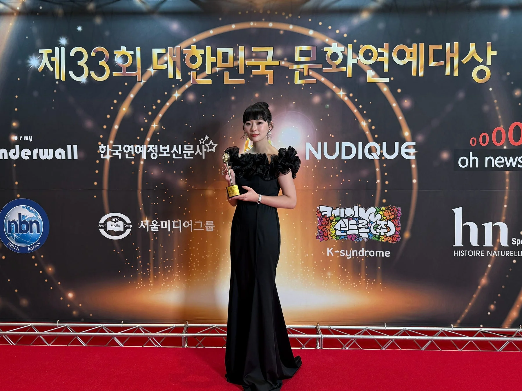 第33回大韓民国文化芸能大賞の授賞式で、黒いドレスをまとった女性がレッドカーペット上でトロフィーを手に笑顔を見せている。背景にはイベント名やスポンサーロゴが輝く。