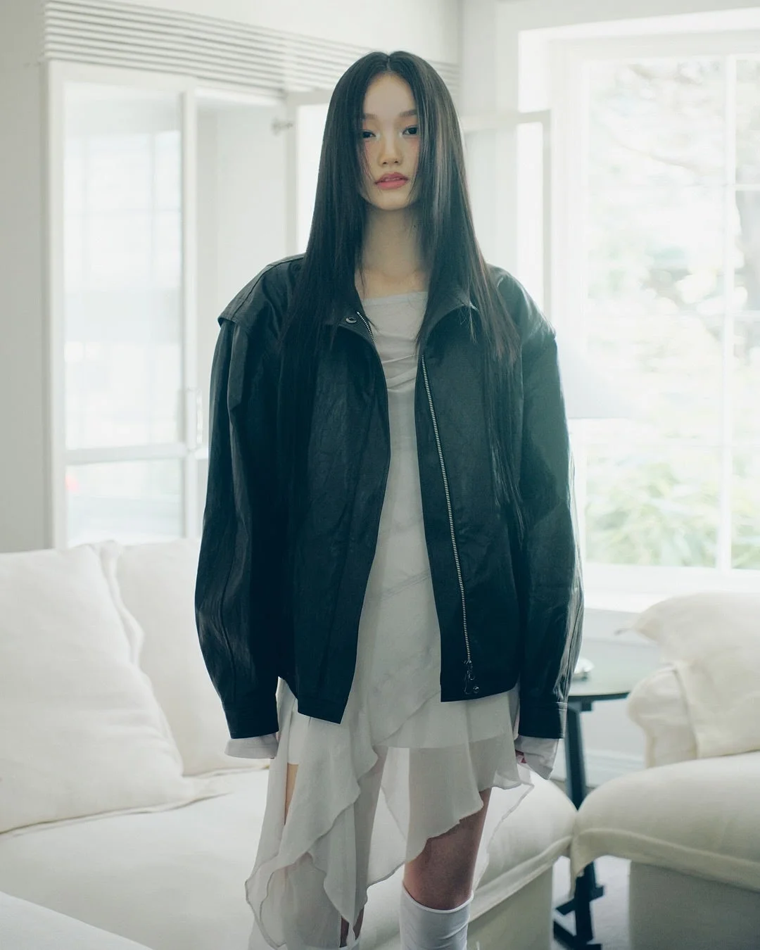 明るい室内の白いソファと窓を背景に、黒いジャケットと白いフリルドレスを着用したロングヘアの女性