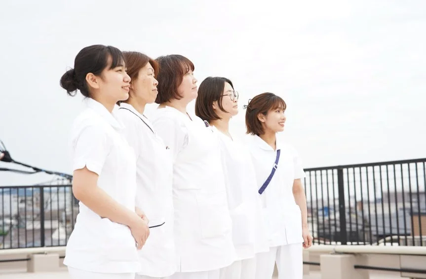 白衣を着た5人の女性たちが、屋外で横一列に並んで同じ方向を見つめている様子