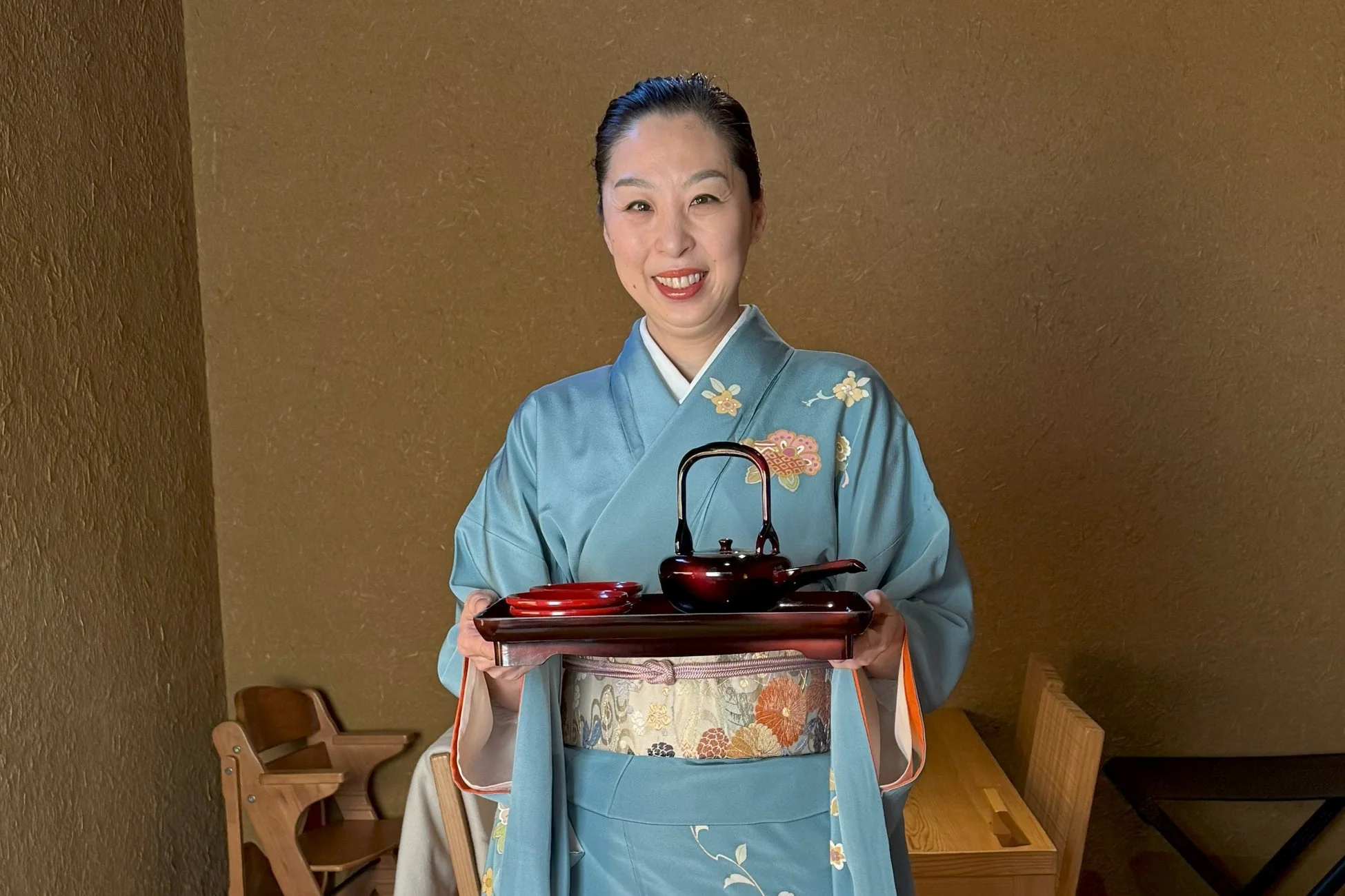 水色の着物を着た女性が笑顔で、茶器と朱色の器が載ったお盆を両手で持っている様子
