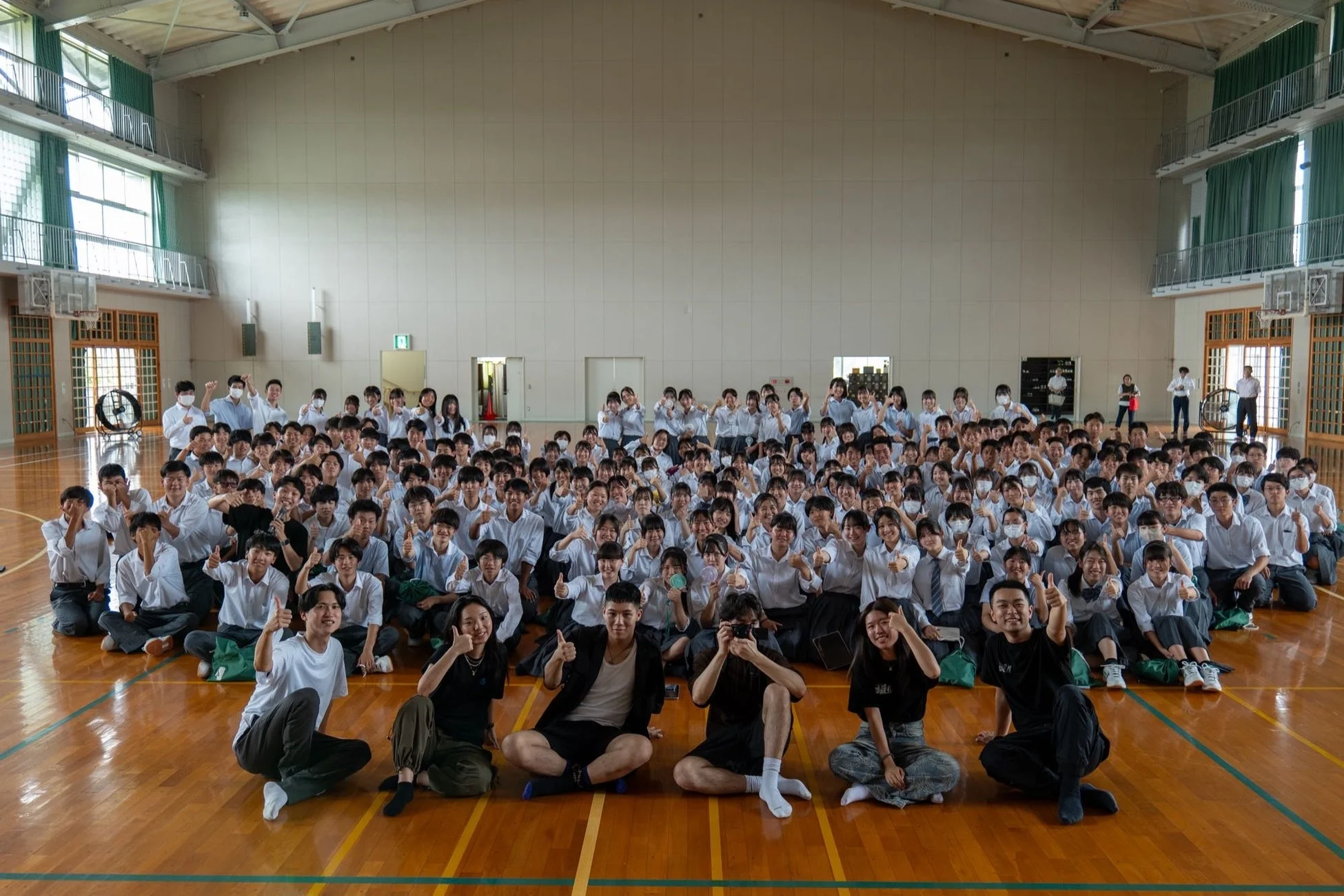 体育館で撮影された、多数の高校生と数名の若者の集合写真。生徒たちは白いシャツの制服を着用し、多くの人が笑顔でサムズアップポーズをとっています。活気ある学校生活の一場面を捉えた一枚です。